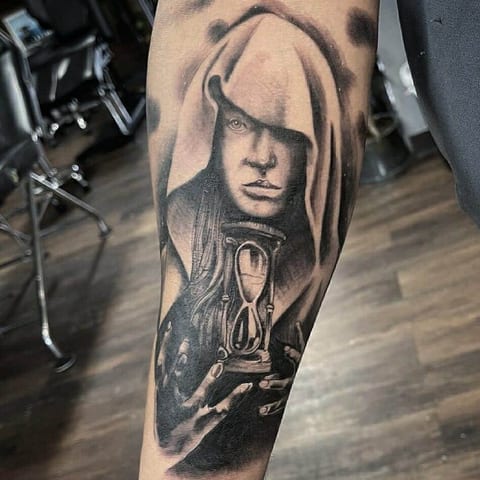 A black and grey realism tattoo of a hooded woman holding an hourglass covers a forearm.
