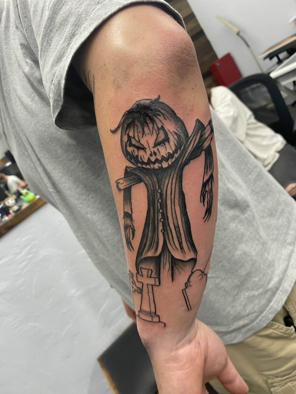 A black and grey pumpkin-headed scarecrow and gravestones cover a forearm.