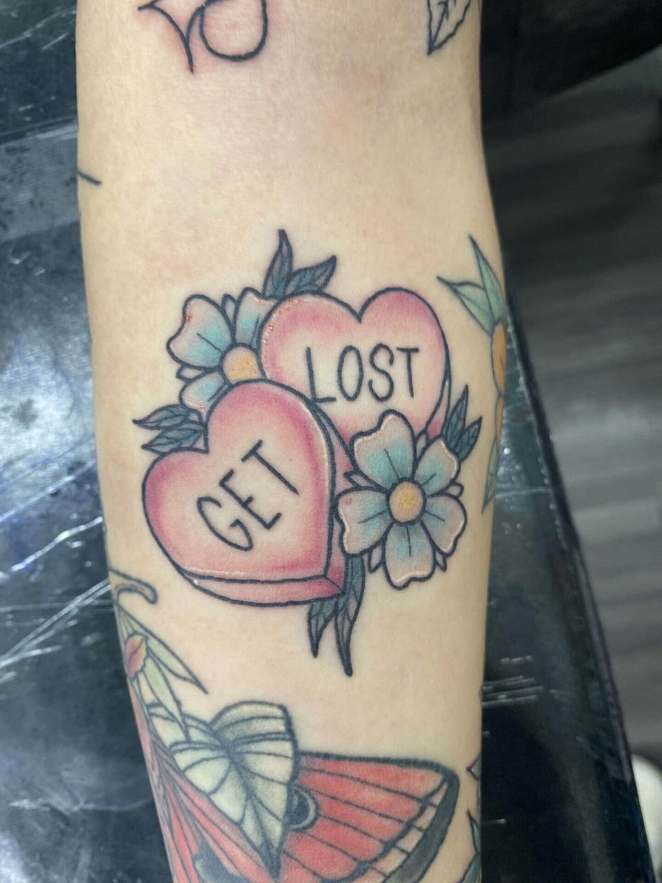 Traditional pink candy hearts reading 'Get lost' sit with blue flowers on a forearm.