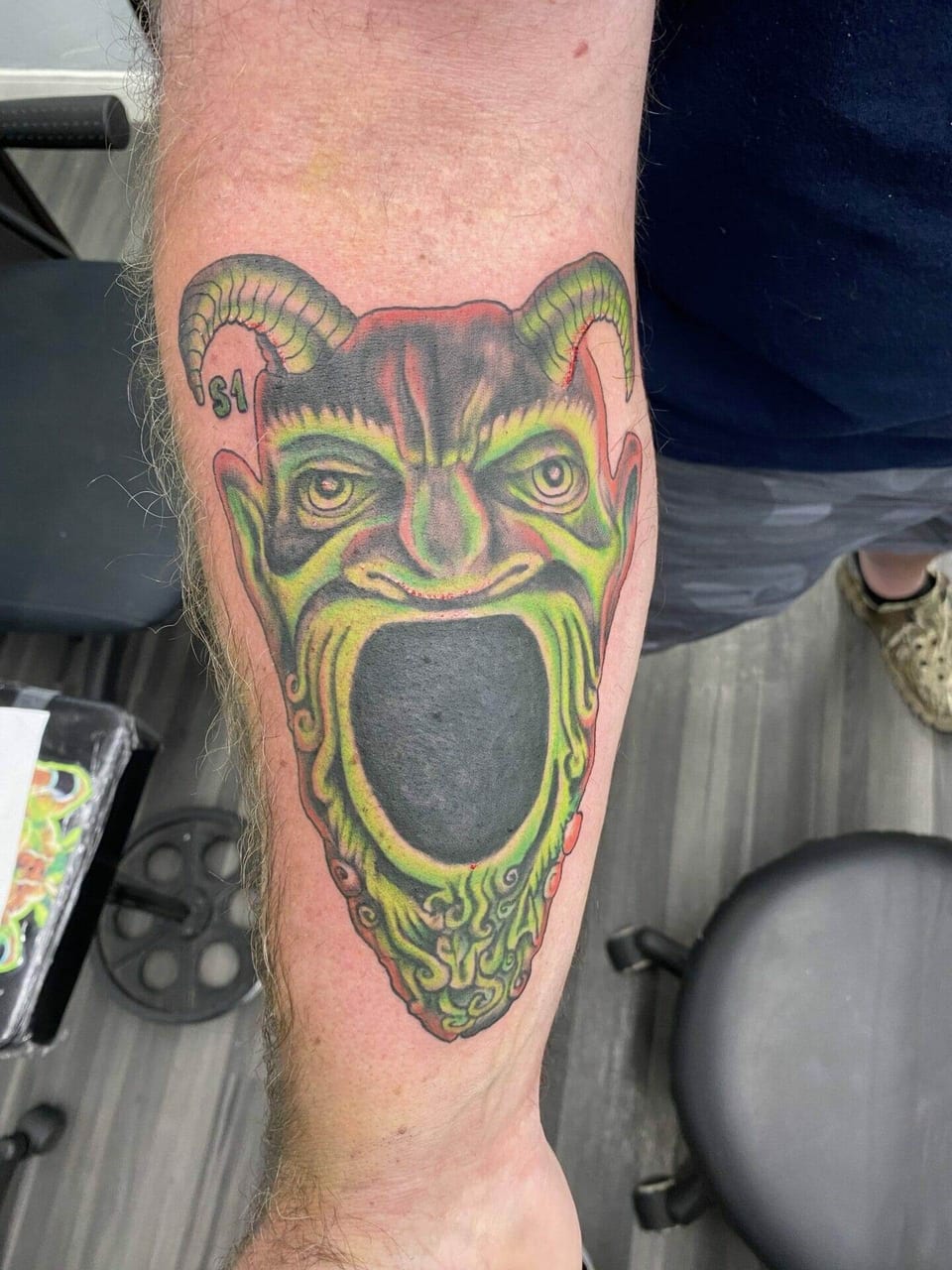A green and red horror tattoo of a horned screaming face covers a forearm.