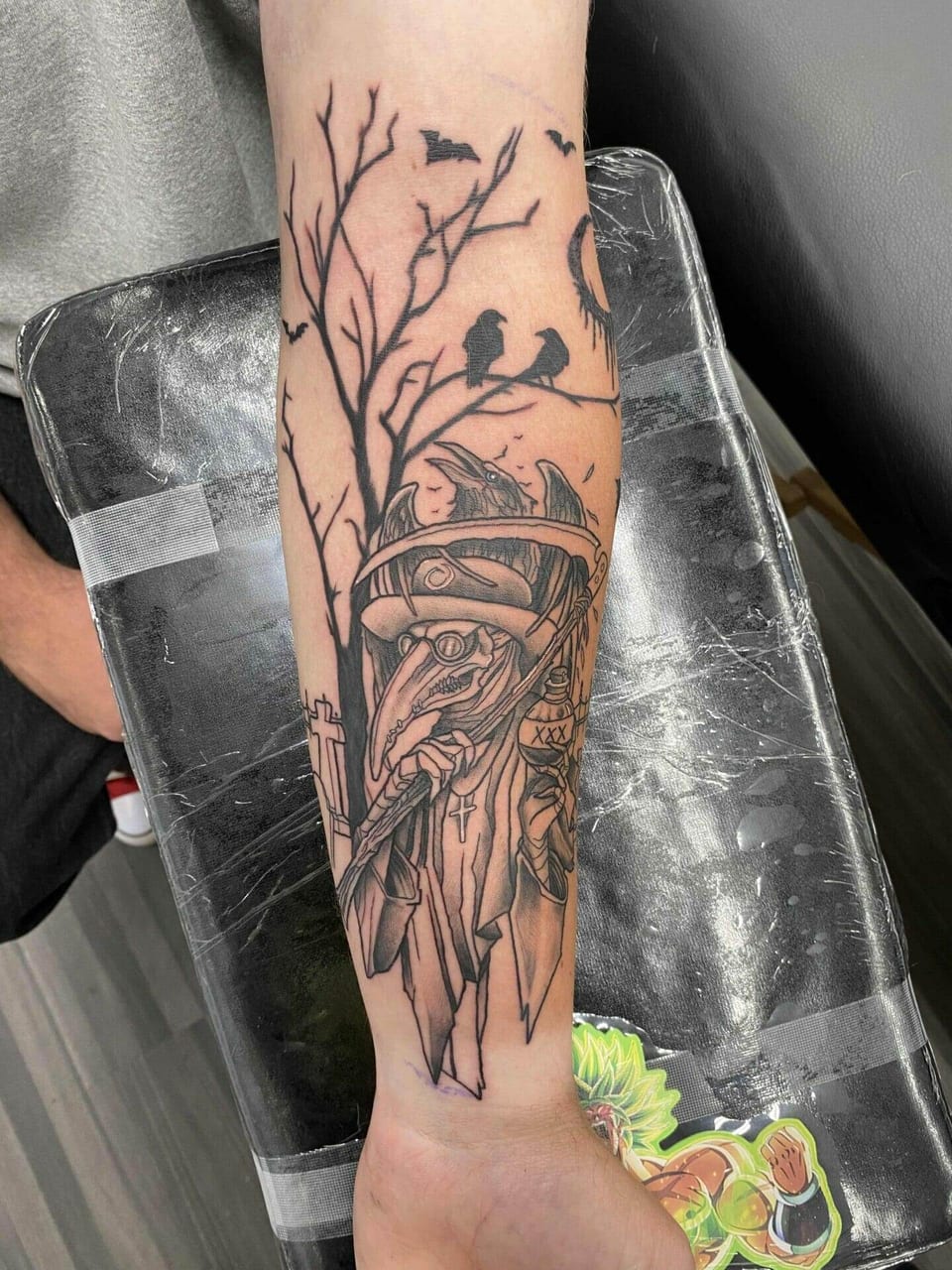 A black illustrative plague doctor, ravens and bare branches cover a forearm.