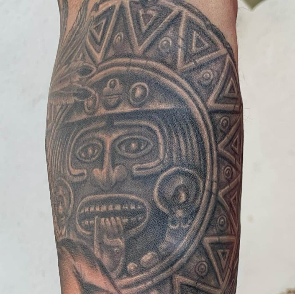 A black and grey Aztec calendar face fills an arm tattoo.