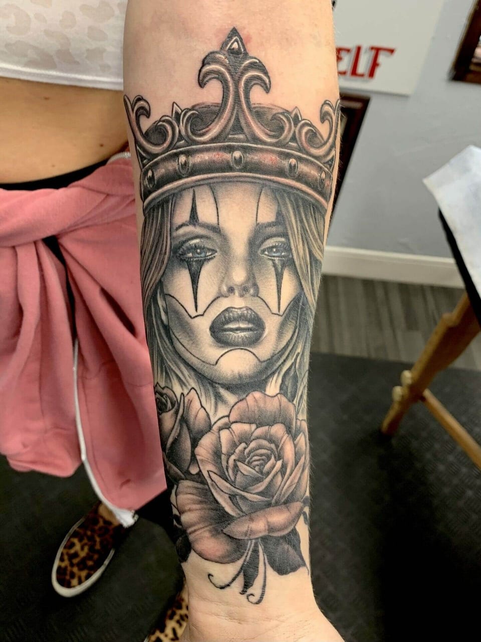 A black and grey crowned woman with clown makeup and roses covers the inner forearm.