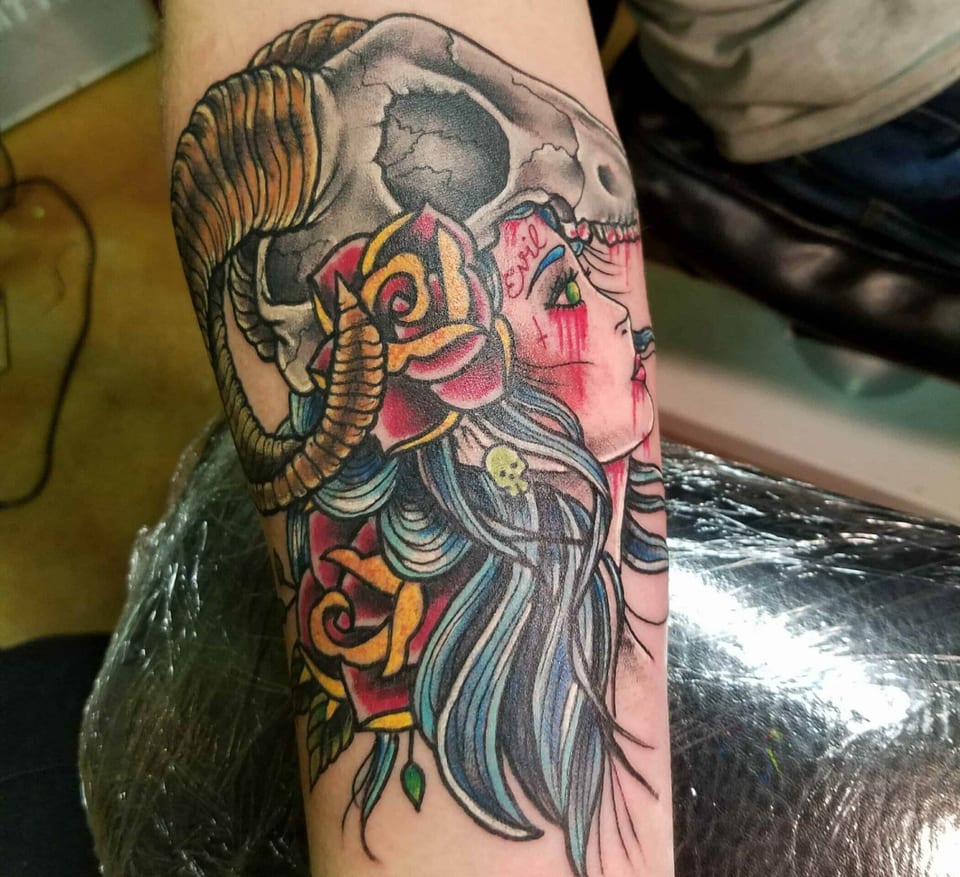 Close view of a ram skull, yellow roses and a neo-traditional woman with red tears and blue hair on a forearm.