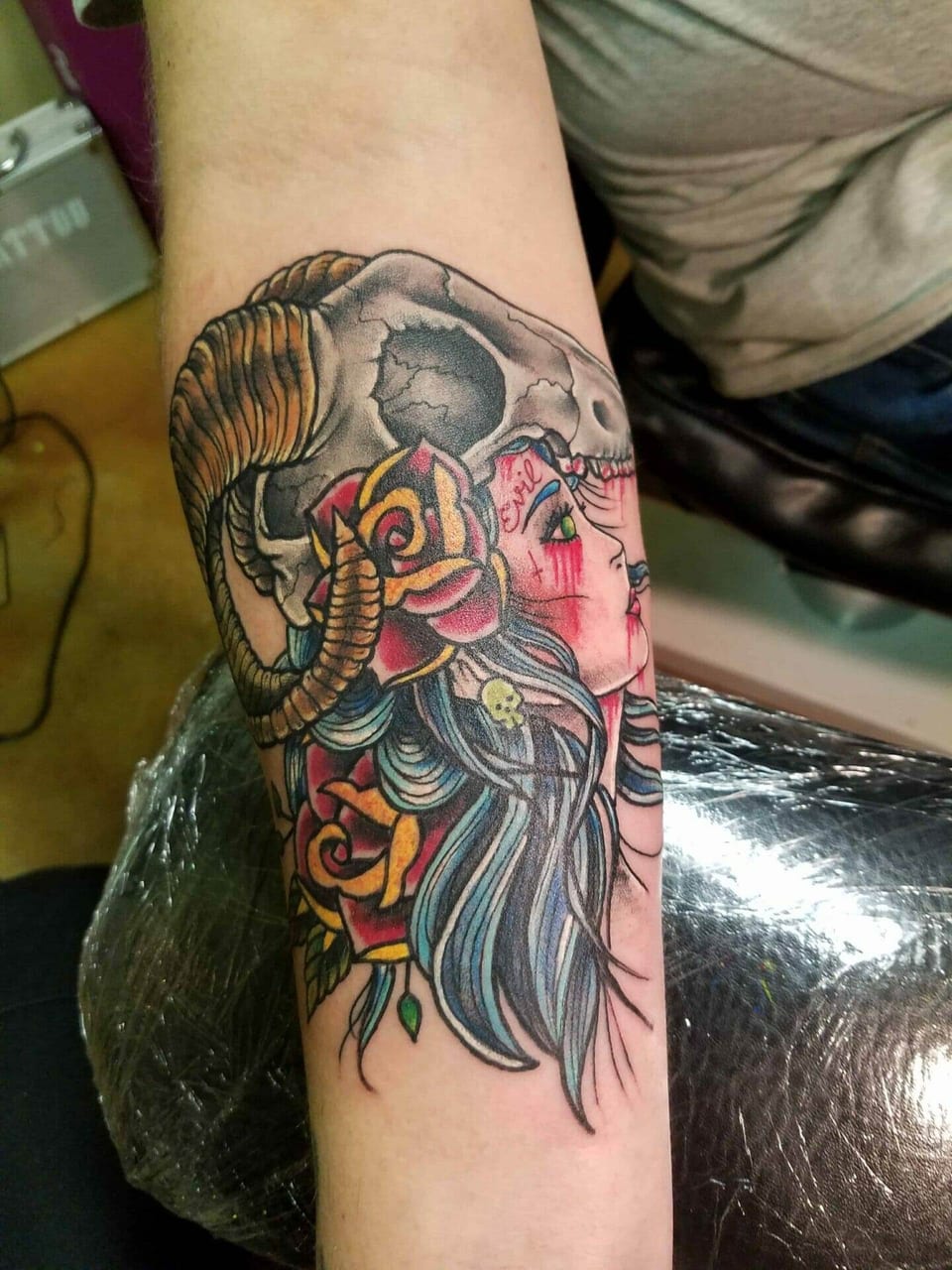 A neo traditional woman wears a ram skull above flowing blue hair and red and yellow roses on a forearm.
