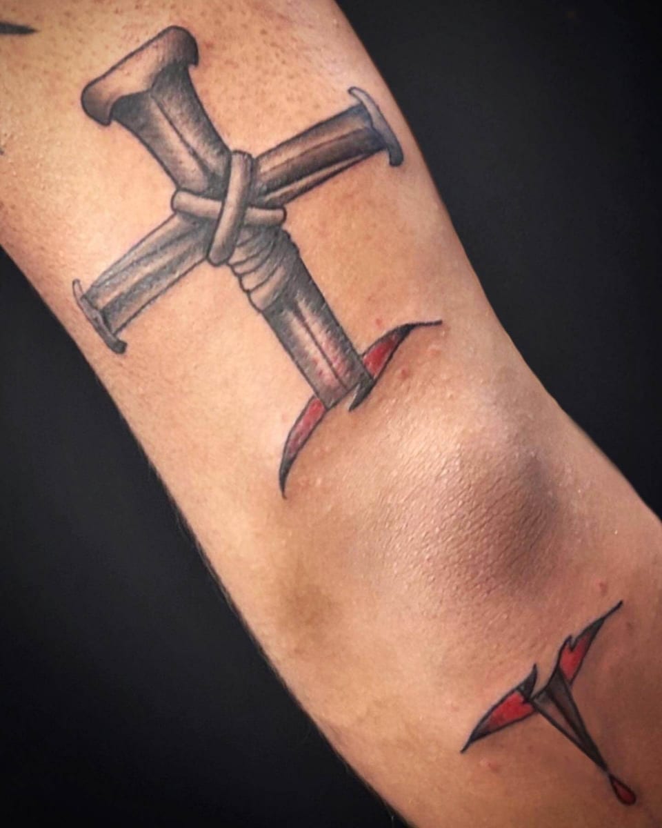 A black and grey cross made of bound nails sits above the inner elbow with small red accents.