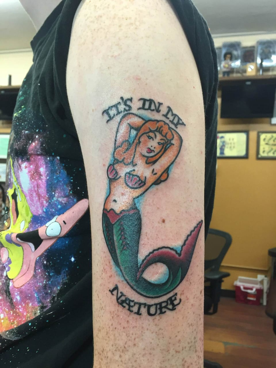 A traditional red-haired mermaid with a green tail sits between curved lettering on the upper arm.