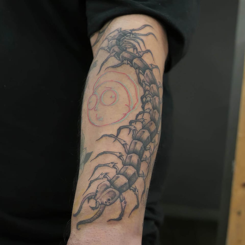 A black and grey centipede curves along the forearm beside a faint colored face outline.