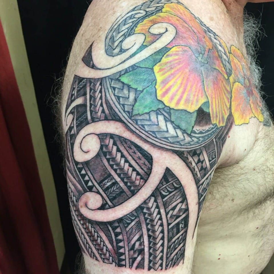 A color hibiscus sits above black Polynesian bands and spirals on a man's shoulder, shown against a dark curtain.