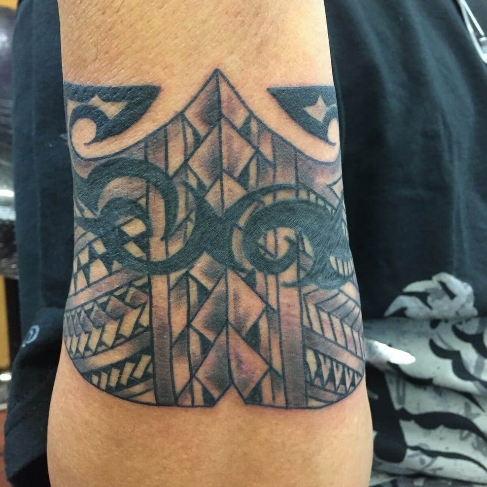 A black Polynesian upper-arm band has interlocking spirals over shaded geometric rows.
