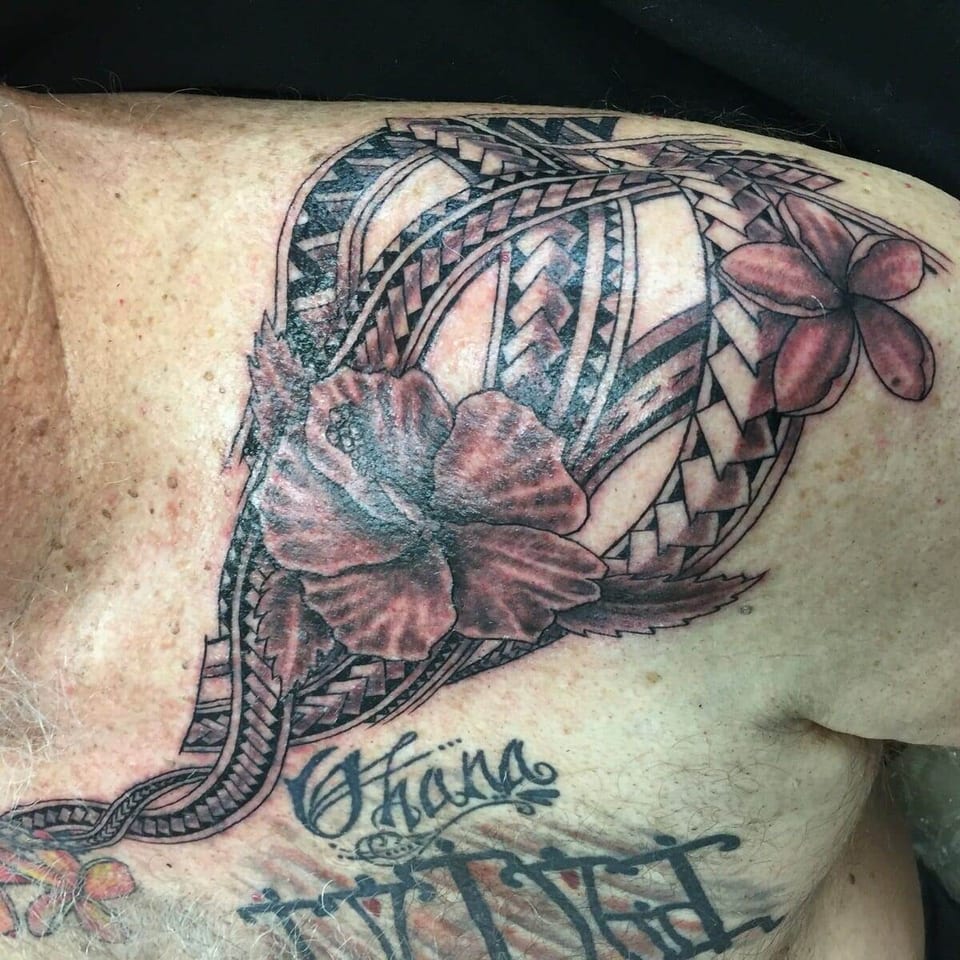 Black and grey hibiscus and plumeria flowers sit among Polynesian geometric bands across the chest and shoulder.