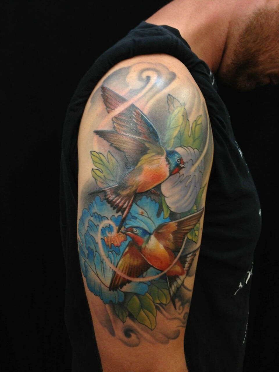 An illustrative color tattoo places flying birds among blue flowers, green leaves and curling clouds on an upper arm.