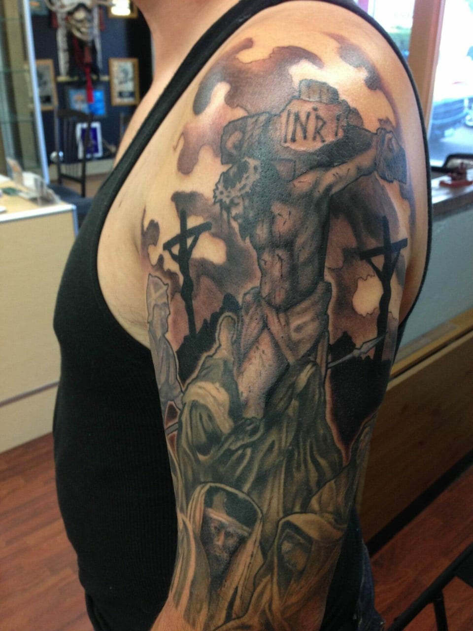 A black and grey crucifixion tattoo fills an upper arm with crosses and clouds.