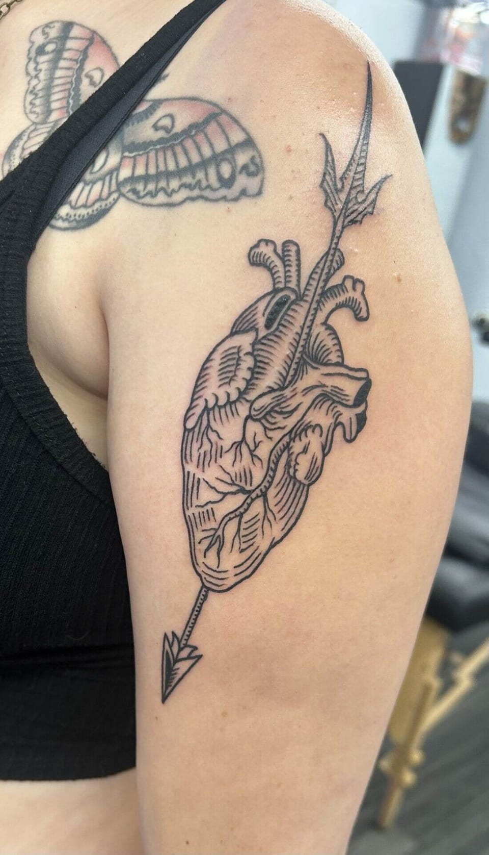 A black linework anatomical heart tattoo is pierced by an arrow on an upper arm.