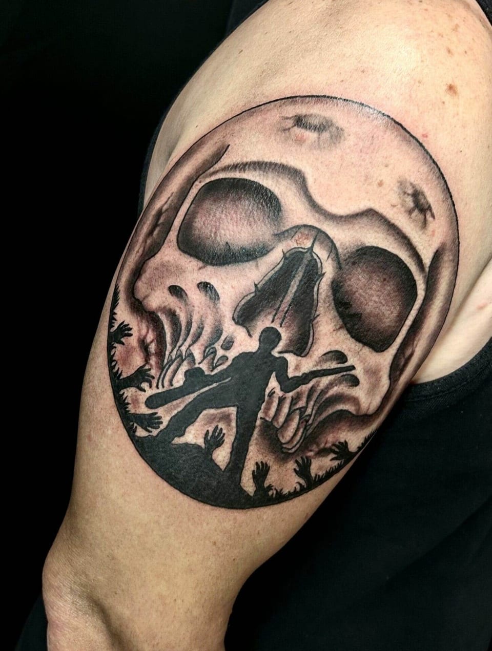 A black and grey skull frames an armed silhouette and reaching hands on the upper arm.
