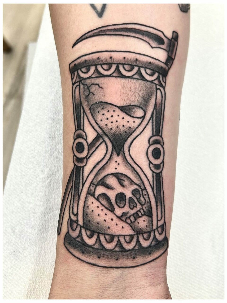 A traditional black and grey hourglass tattoo contains a skull beside a scythe on a forearm.