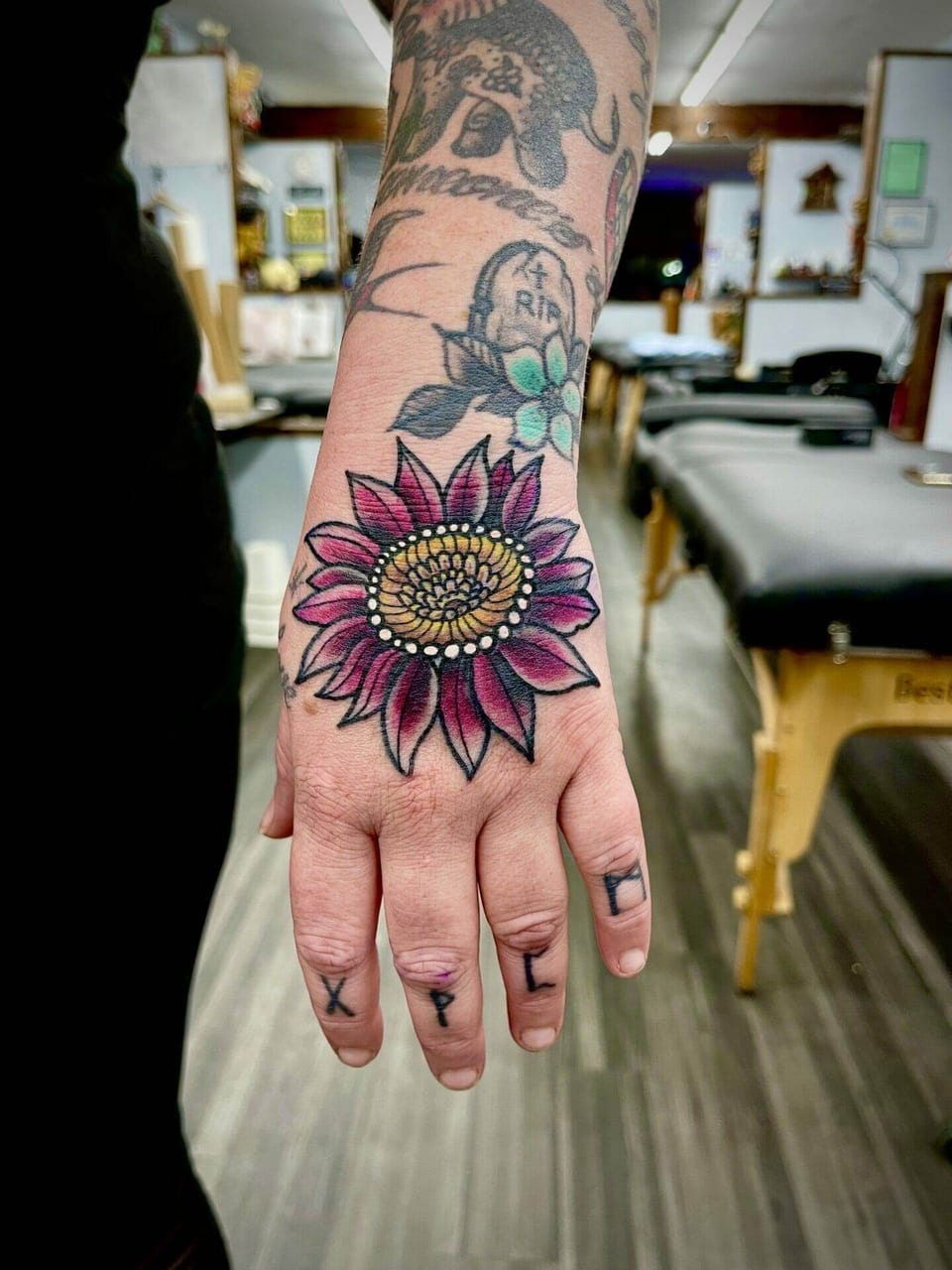 A neo traditional purple sunflower with a golden center covers the back of a hand.