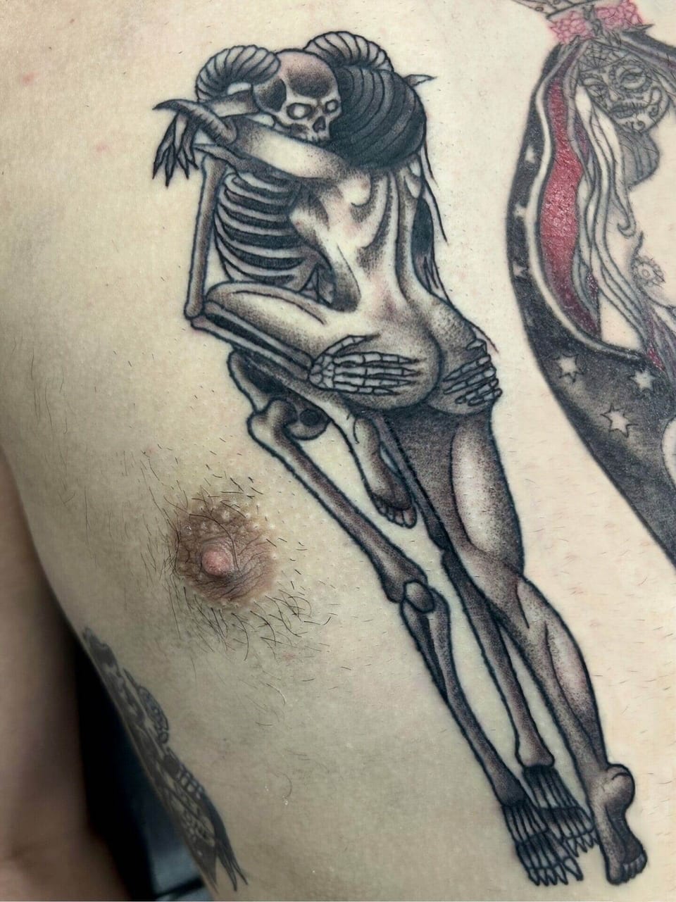 A black and grey illustrative tattoo shows a woman embracing a skeleton across the chest.