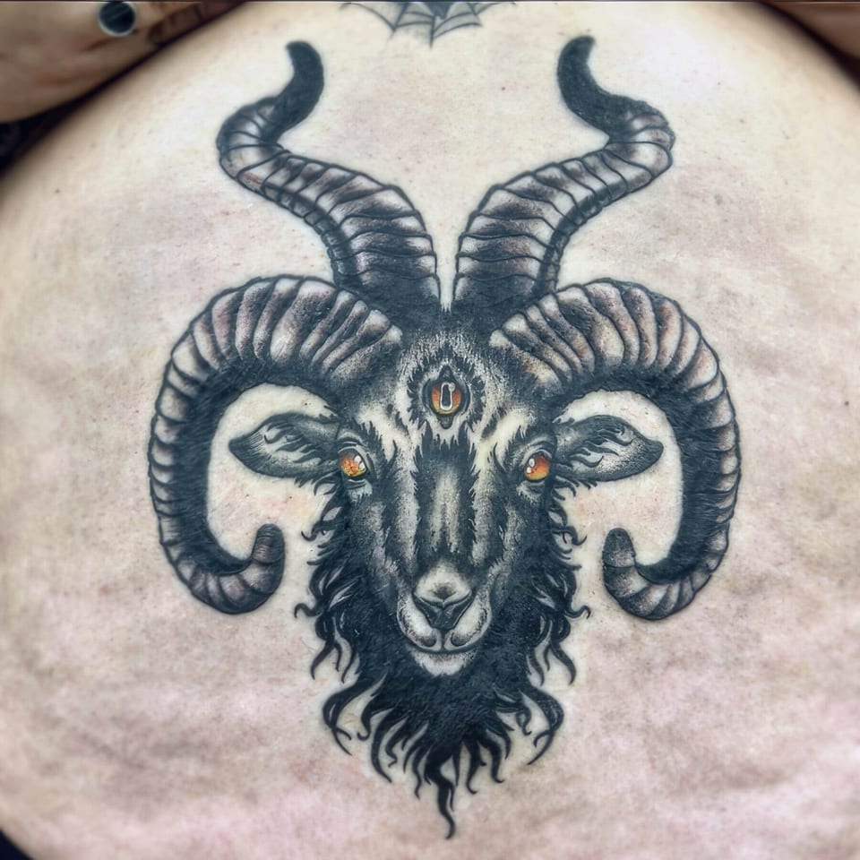 A black and grey goat tattoo with curling horns and orange eyes covers the torso.