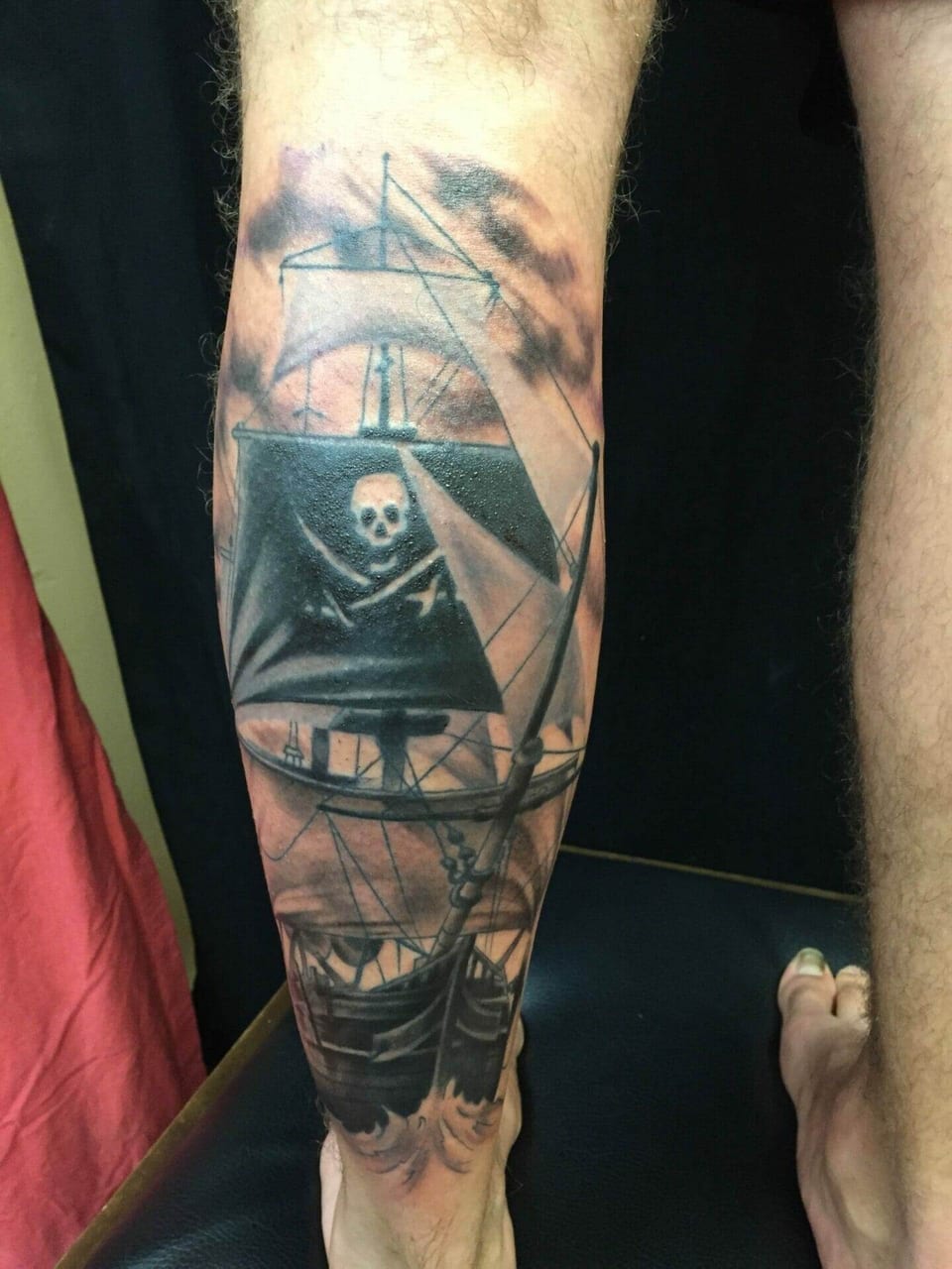 A black and grey pirate ship tattoo carries a skull-and-crossbones sail down a calf.