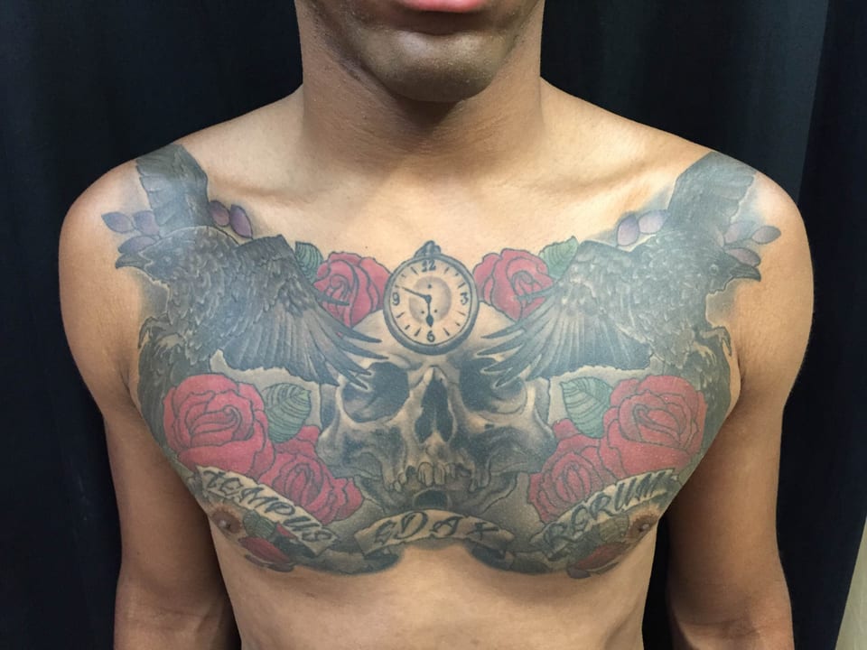 A traditional chest tattoo combines a skull, birds, red roses, pocket watch and lettering.
