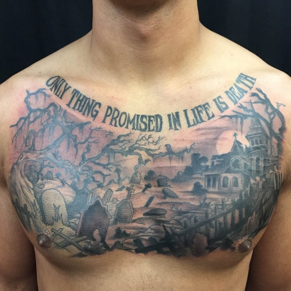 A black and grey chest tattoo shows a graveyard, bare trees and church beneath lettering.