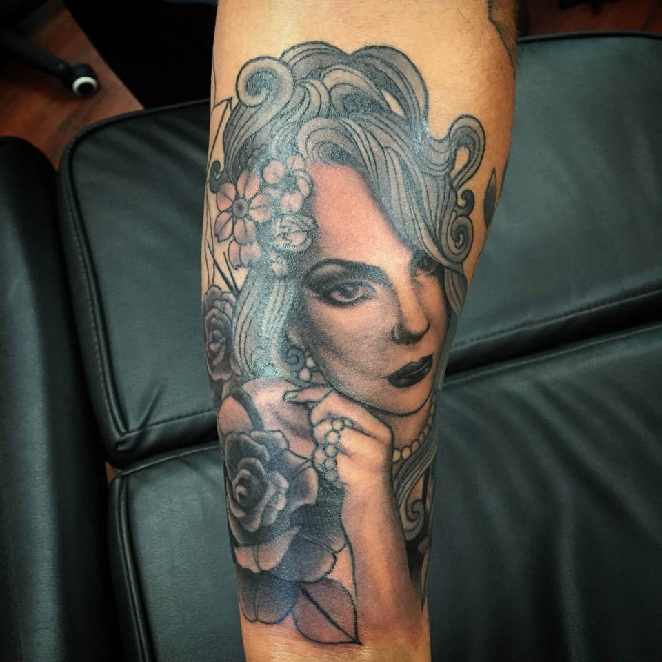 An illustrative black and grey woman tattoo wears flowers and pearls beside roses on a forearm.
