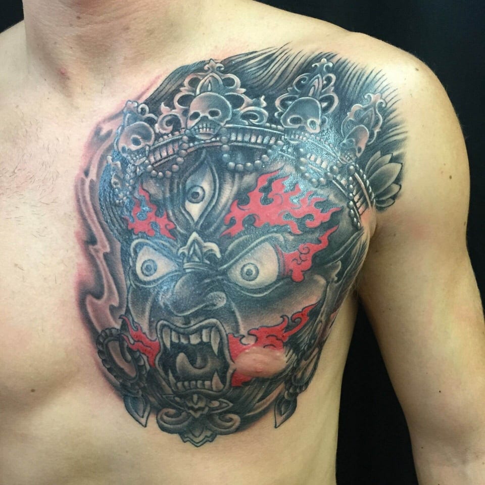 A black and grey demon mask tattoo has a skull crown and red flames across the chest.
