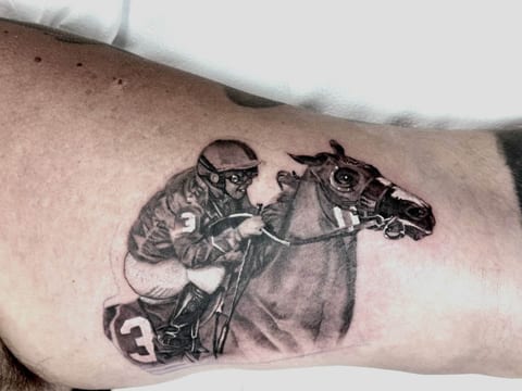 A black and grey jockey leans forward over a racehorse on an upper arm.