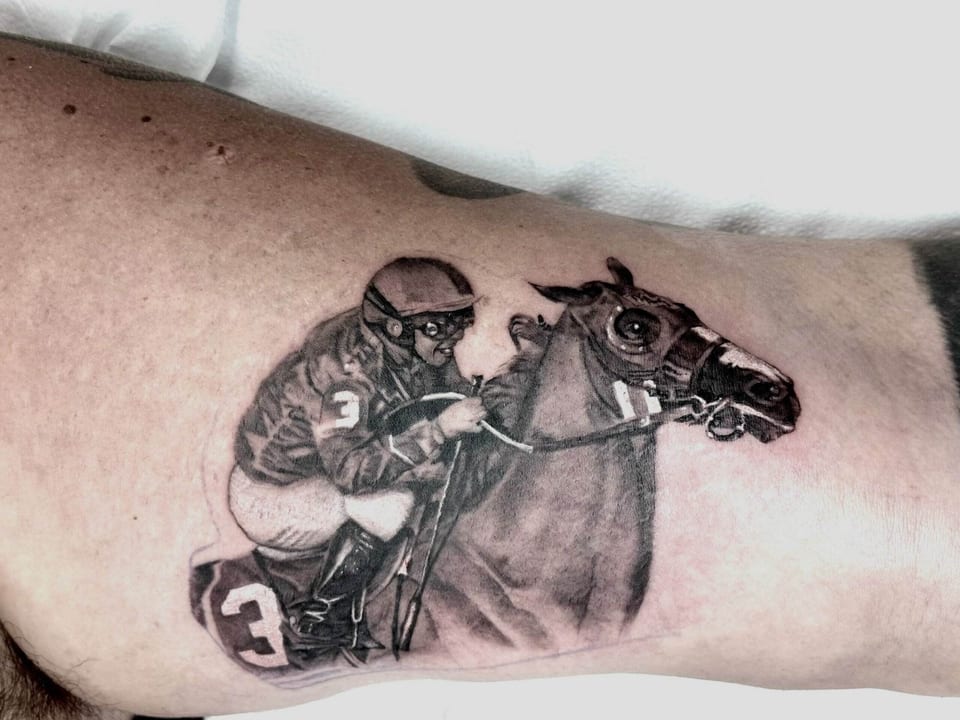 A black and grey jockey leans forward over a racehorse on an upper arm.