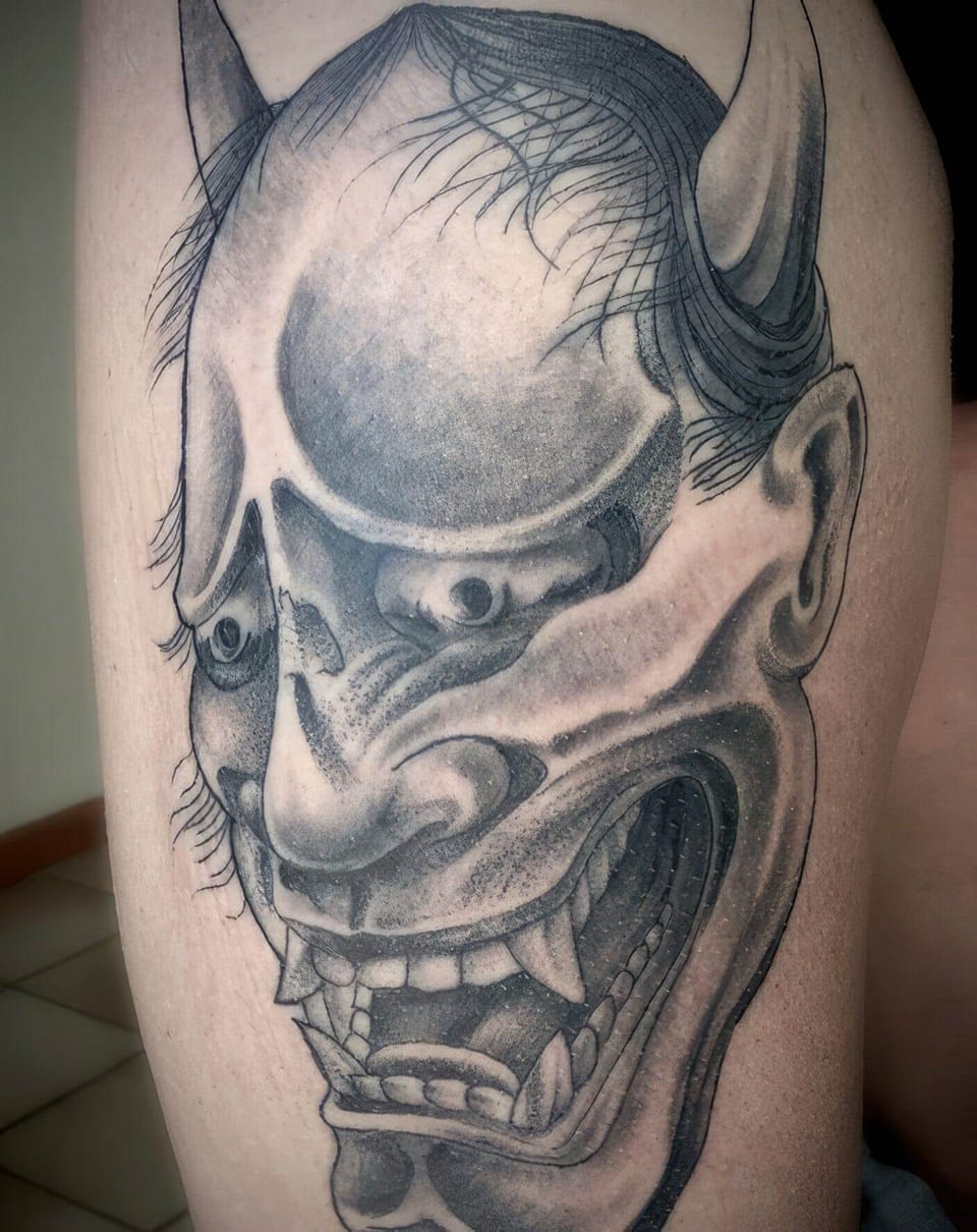 A black and grey Japanese demon mask with horns and fangs covers a thigh.