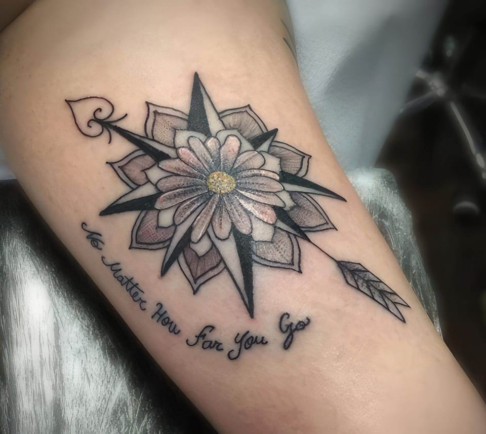 A flower with a yellow centre sits inside a compass pierced by an arrow above script on a forearm.
