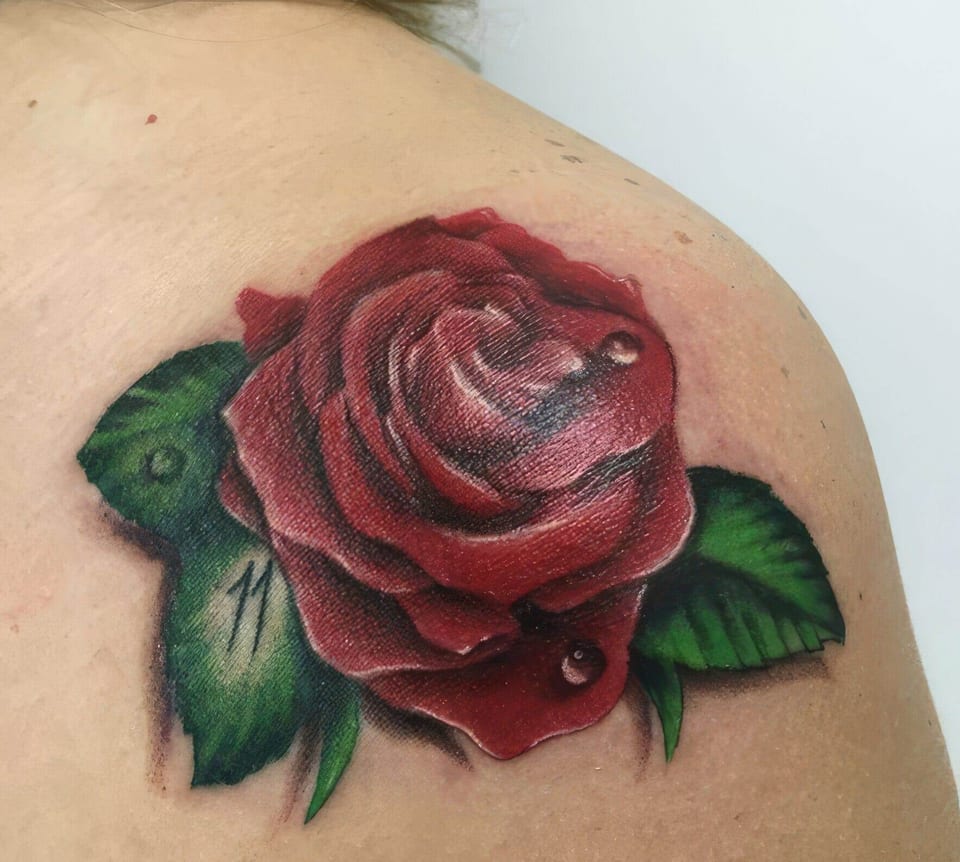 A realistic red rose with green leaves and water droplets sits on the shoulder blade.