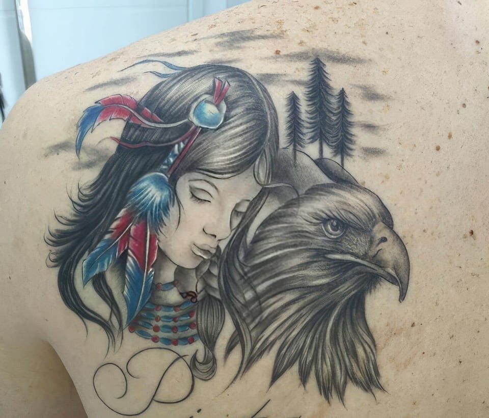 A neo traditional woman with red and blue hair feathers sits beside an eagle and pine trees on the shoulder blade.