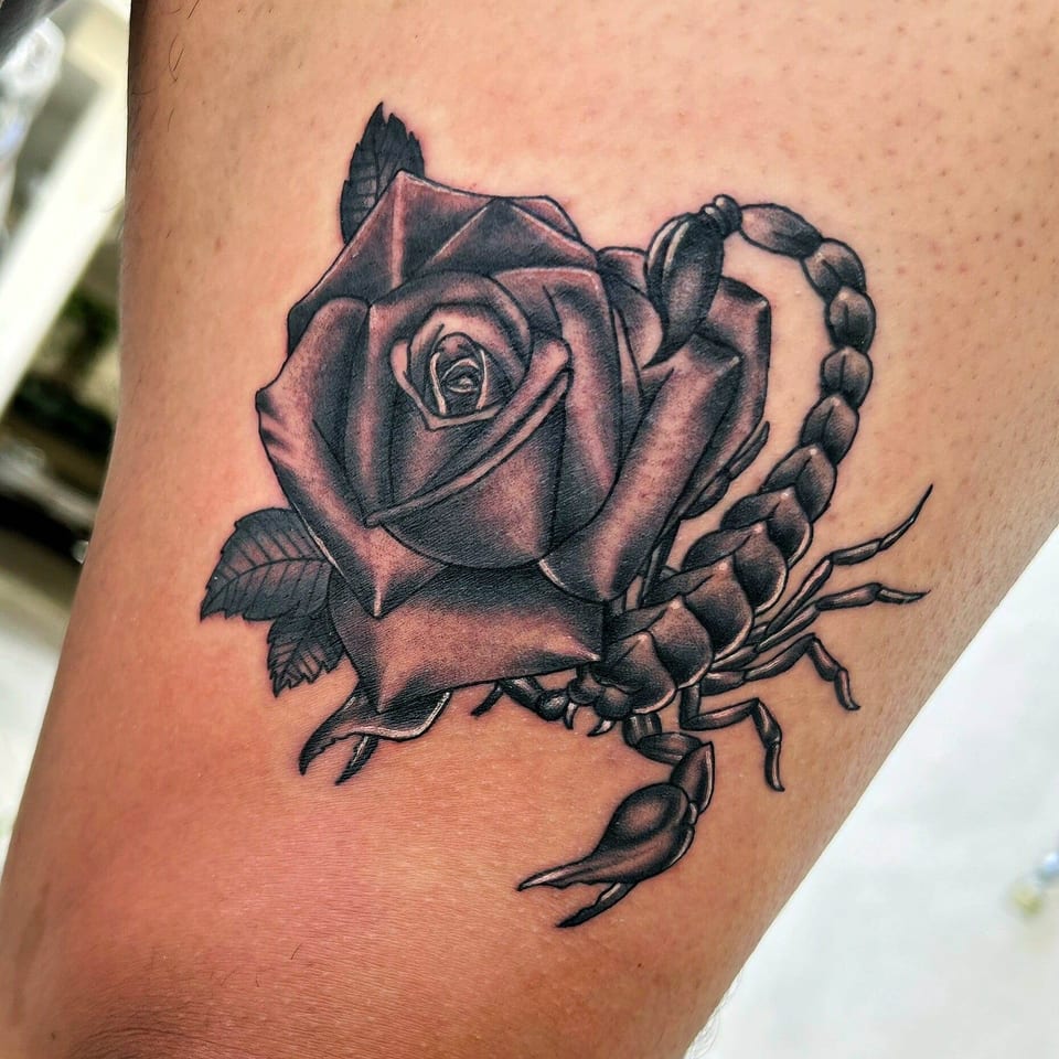 A black and grey scorpion curves around a shaded rose on a thigh.