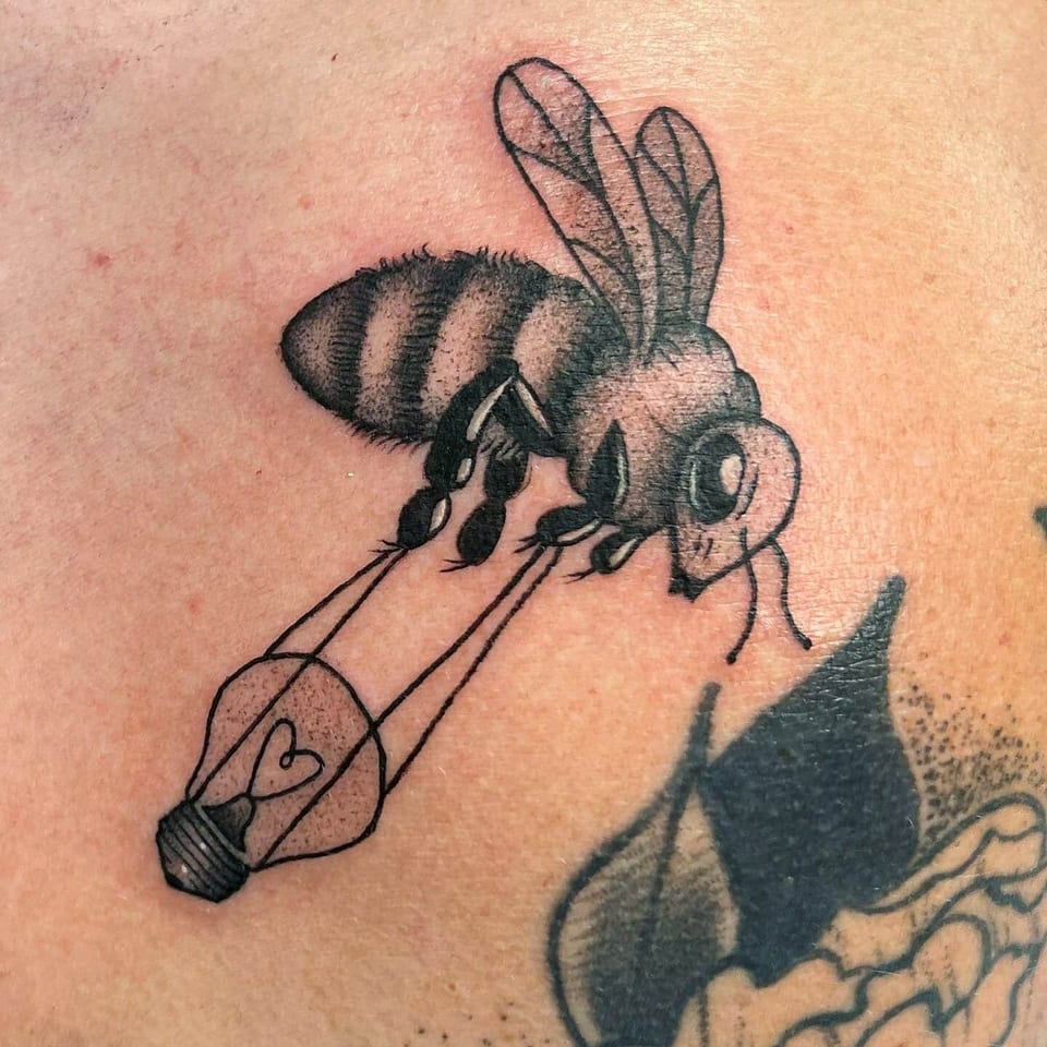 A black and grey bee carries a light bulb with a heart-shaped filament on an arm.