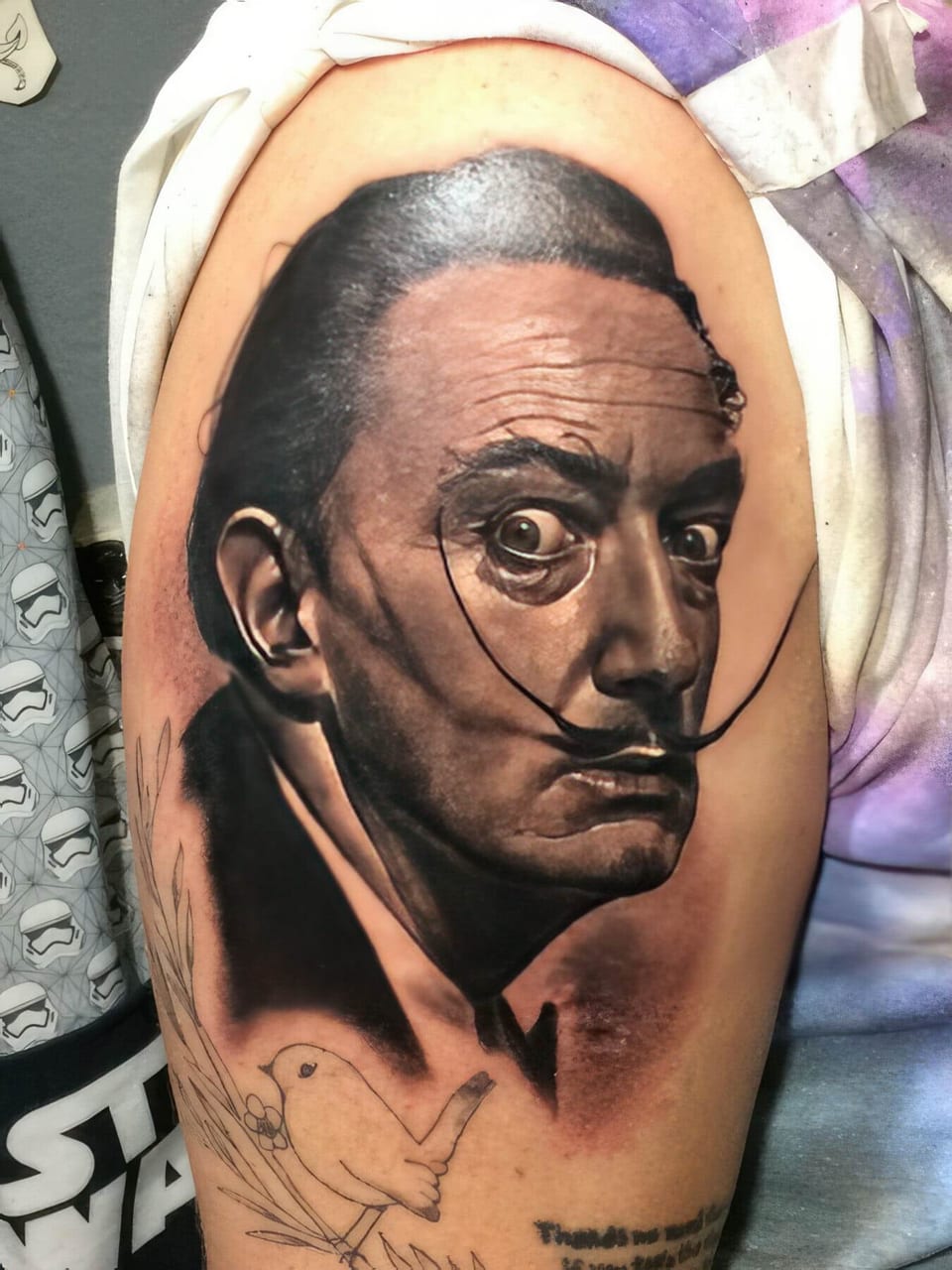 A realistic portrait tattoo of a man with an upturned moustache covers an upper arm.