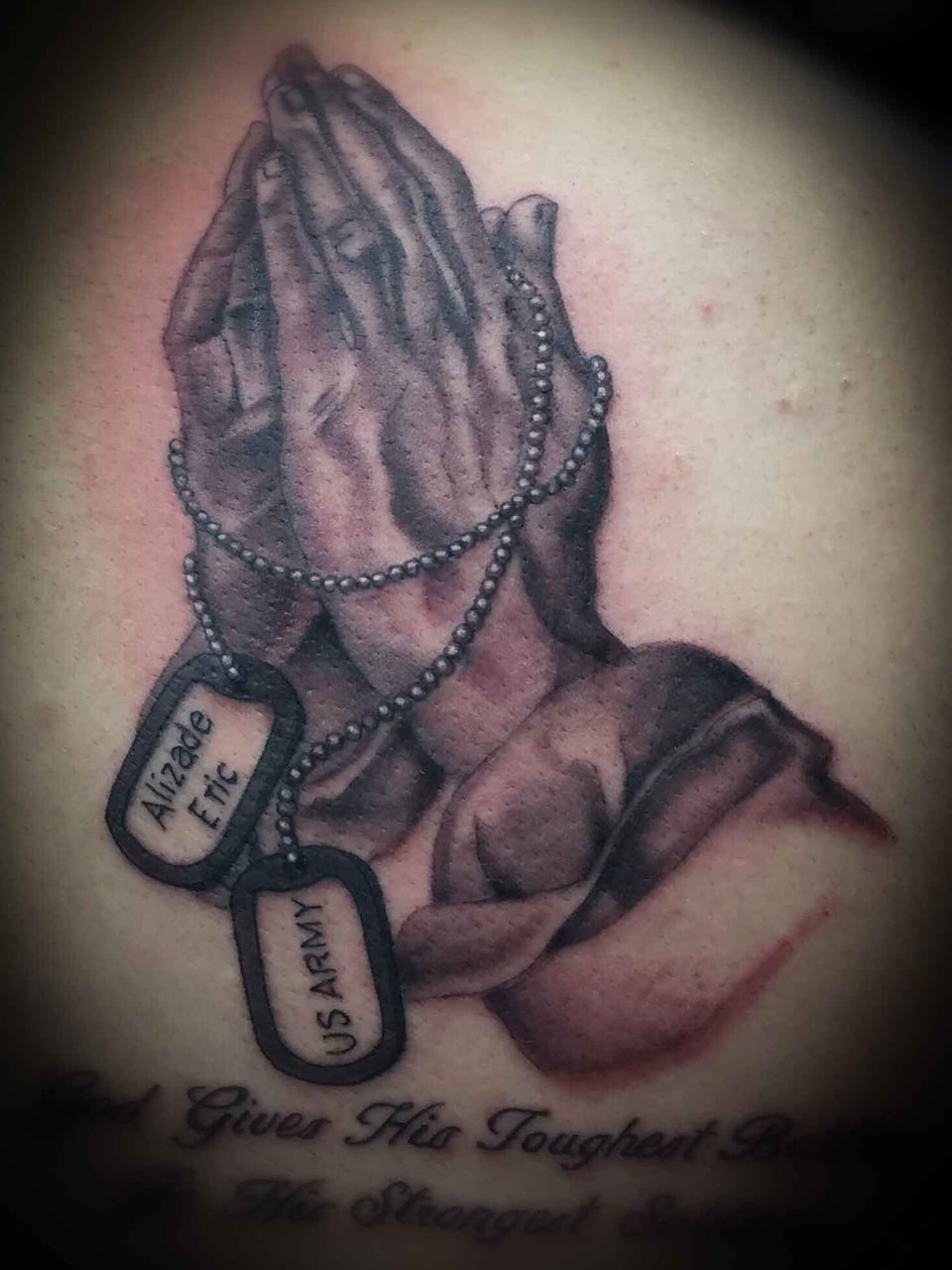 A black and grey tattoo shows praying hands wrapped in beads beside dog tags.