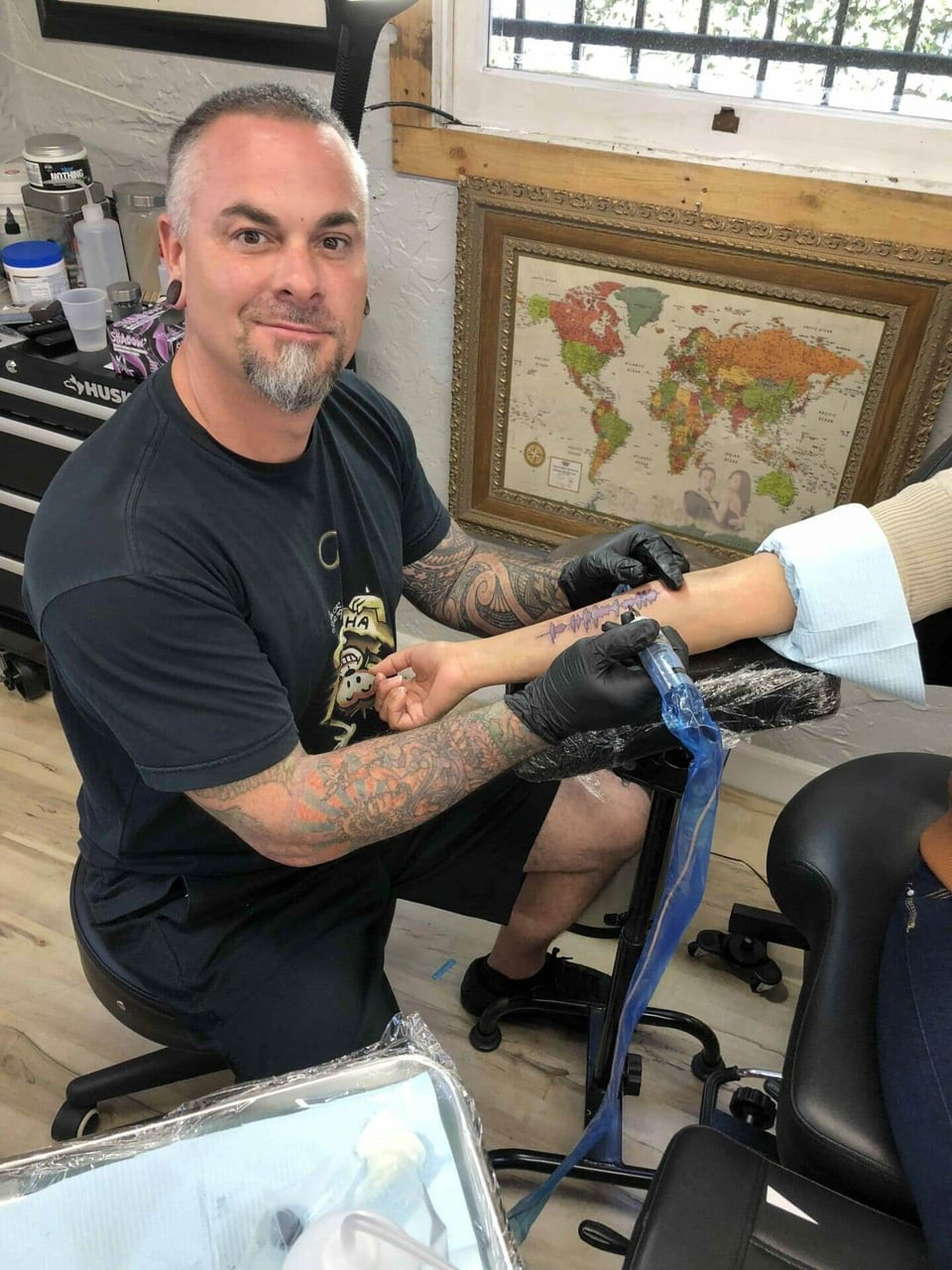 Seth Reynolds tattooing a client's arm beside a framed world map at his station