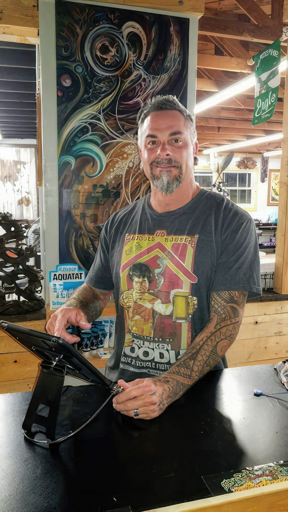 Seth Reynolds at the Funhouse counter, holding a tablet with his tattooed arms in front of the mural