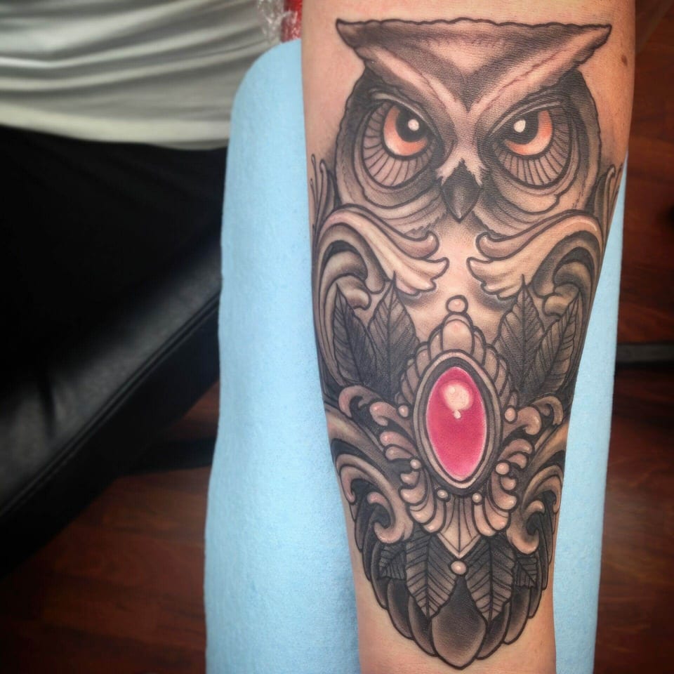 A neo-traditional shaded owl with orange eyes and an ornamental pink gem covers the forearm.