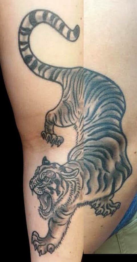 A shaded tiger prowls down an inner upper arm with its mouth open and tail curled.