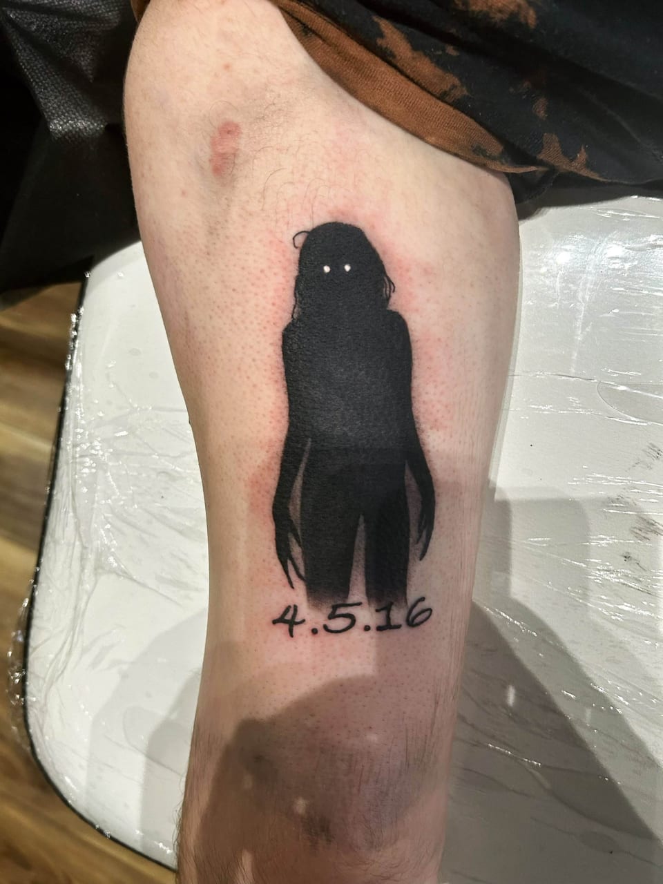 A black silhouette of a person with white eyes is tattooed on an arm above a date The tattooed arm is resting on a white surface.