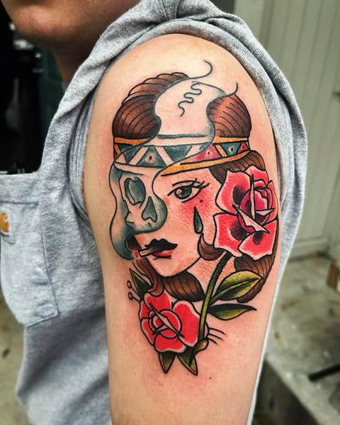 A colorful tattoo on an upper arm depicting a woman's face with a headdress, surrounded by roses and leaves, and partially covered by a skull design.