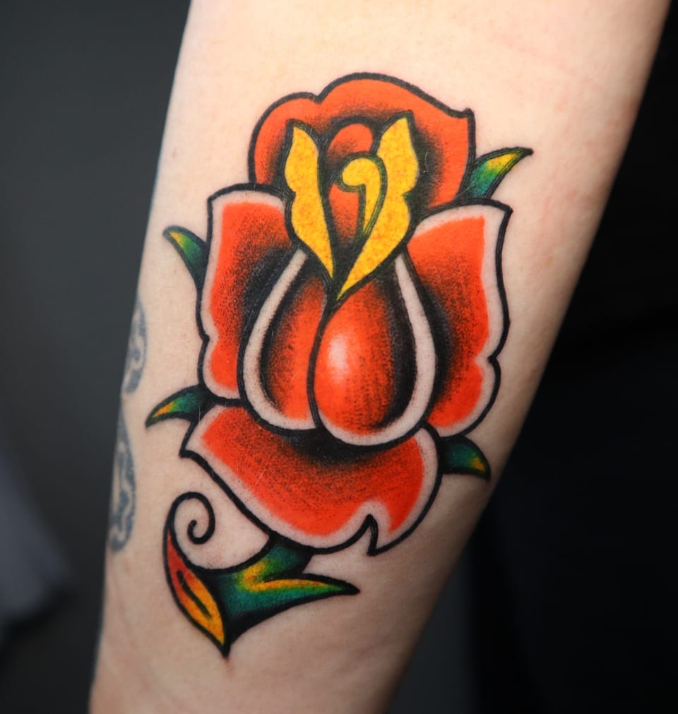 A red traditional rose tattoo opens around a yellow center and green leaves.