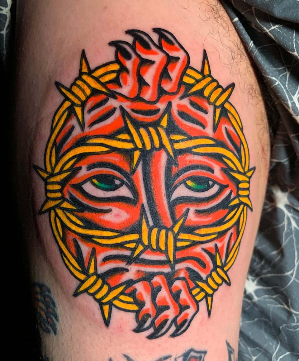 A red clawed face tattoo peers through a yellow barbed-wire ring.