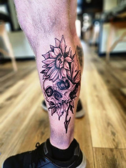Black and grey illustrative skull and sunflower tattoo on a calf.