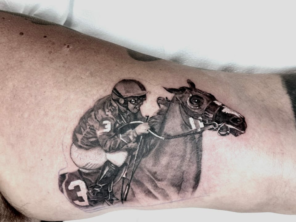 A black and grey realism jockey riding a horse covers an inner upper arm.