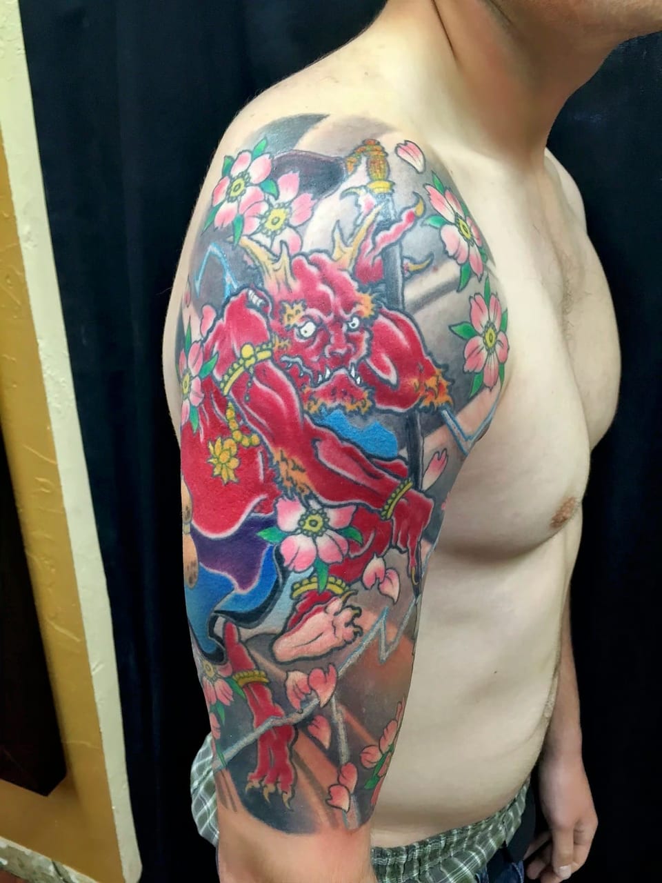 Japanese tattoo of a red demon and pink blossoms covers the upper arm and shoulder.