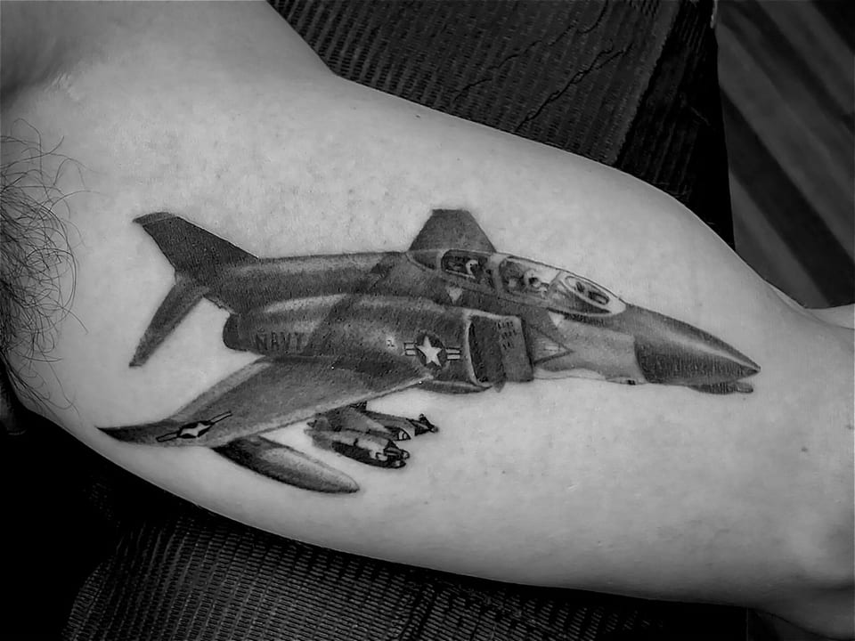 Black and grey realism tattoo of a fighter jet spans the inner upper arm.